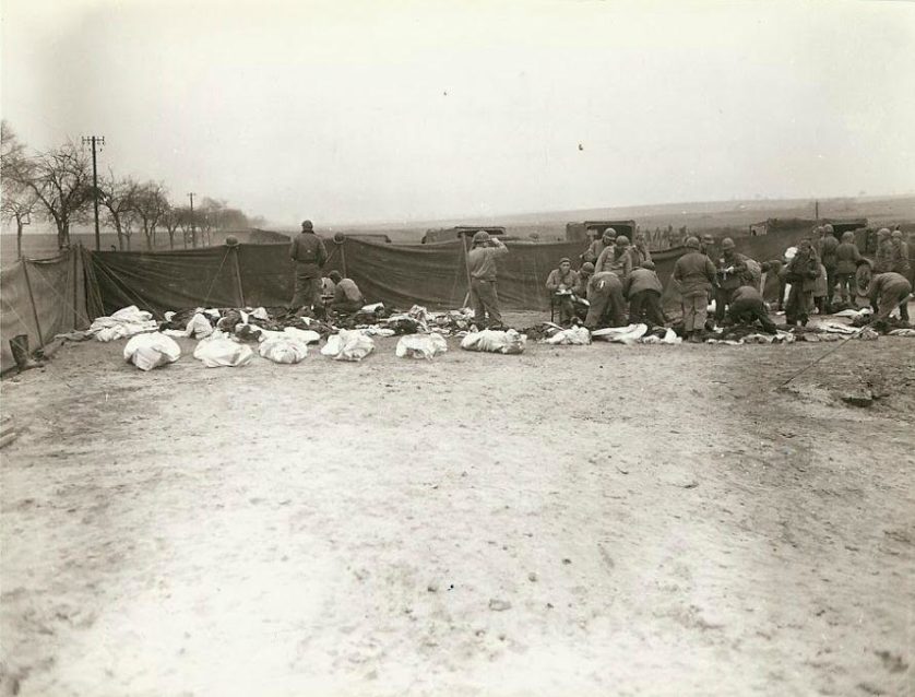 Soldados norte-americanos e alemães mortos em um cemitério antes do enterro, em um local desconhecido. Cada corpo foi colocado em uma capa de colchão. Prisioneiros alemães podem ser vistos fazendo o trabalho de cavar as sepulturas e colocando os corpos dentro delas. 
